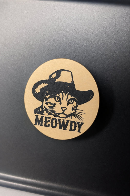 A round gold magnet featuring a black cat face wearing a cowboy hat with the word 'MEOWDY' underneath, photographed on a dark gray surface. 