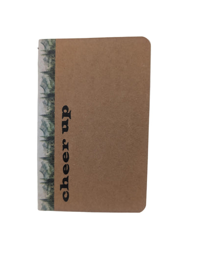 A small kraft paper notebook with a vertical forest patterned spine and the words 'cheer up' printed in black on the front, photographed on a plain light background.