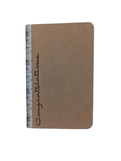 A small kraft paper notebook or journal with rounded corners, featuring the word 'Congratulations' written vertically in black script along the left edge.