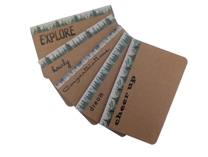 A fan of kraft paper cards with mountain tree patterned borders and motivational words like EXPLORE, howdy, congratulations, dream, and cheer up, displayed on a white background.