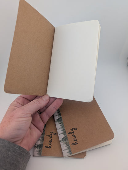 A hand holds an open kraft paper notebook showing blank white pages. Two closed matching notebooks with a forest-patterned band labeled 'howdy' rest on a white surface below.