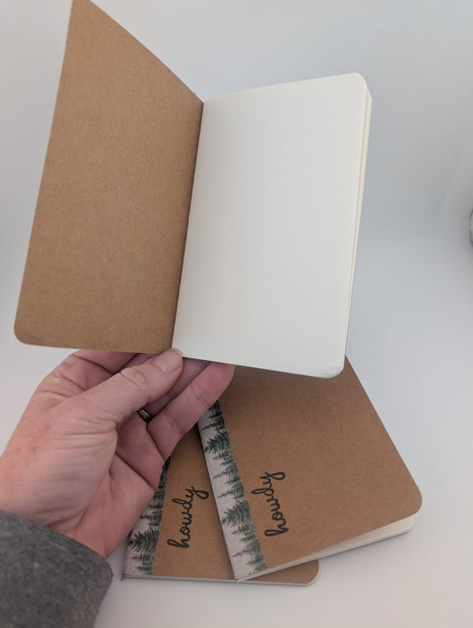 A hand holds an open kraft paper notebook showing blank white pages. Two closed matching notebooks with a forest-patterned band labeled 'howdy' rest on a white surface below.
