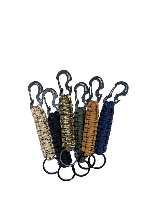 Five paracord keychains in different colors (beige, black, green, brown, and navy) with black carabiners and key rings, arranged in a fan shape on a white background.